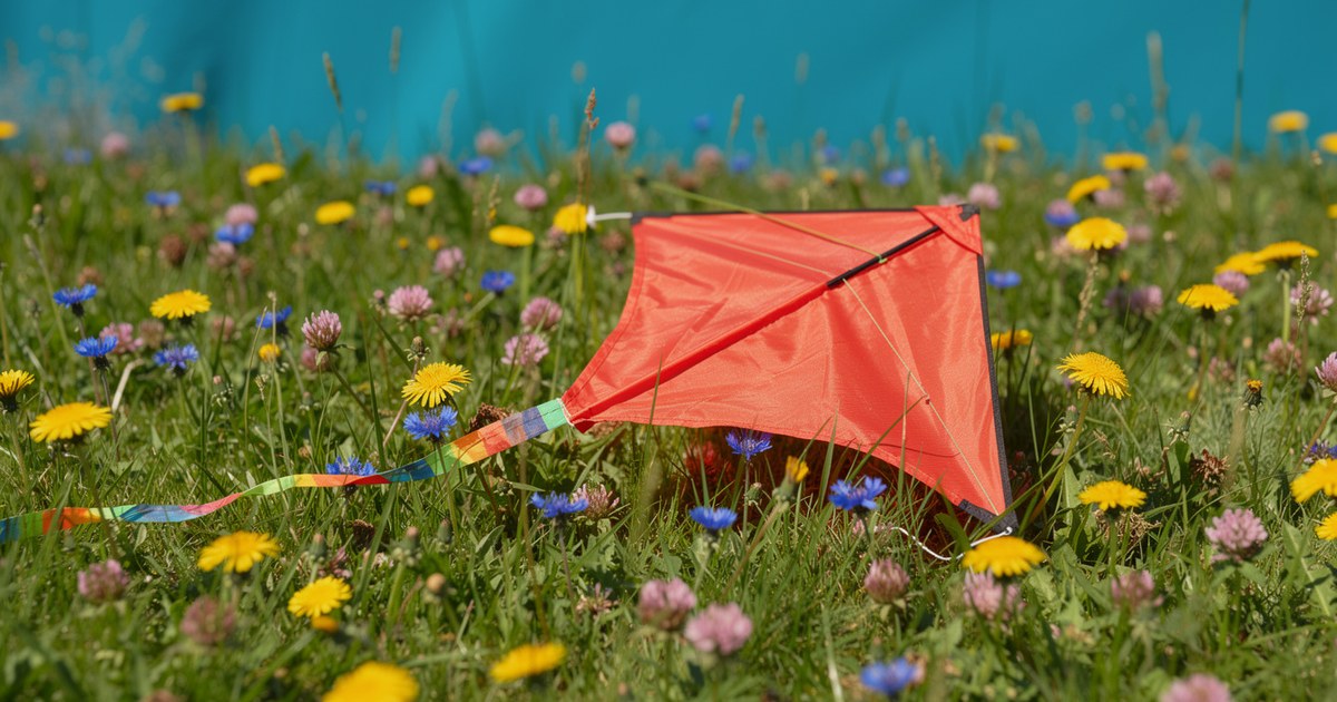 A coral red kite with a rainbow tail resting in a green meadow full of yellow dandelions, blue cornflowers and pink clover.